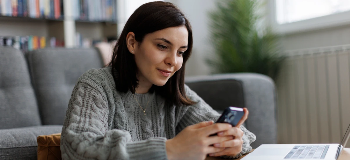 Mulher jovem usando smartphone enquanto trabalha ou estuda em casa, com fundo de estantes de livros e uma planta.