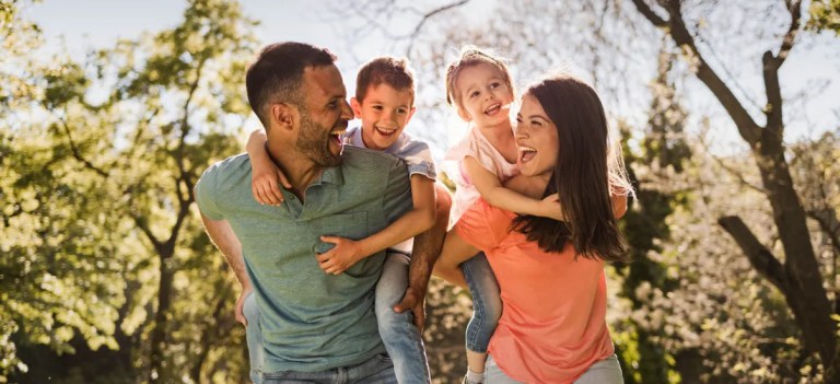 Família sorridente ao ar livre em um parque: pai abraça o filho e mãe segura a filha em meio às árvores, em dia ensolarado.
