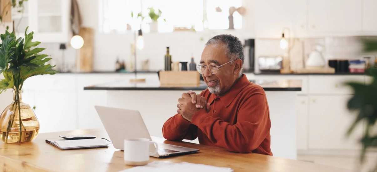 Homem idoso sorrindo enquanto participa de uma reunião virtual no ambiente de cozinha moderna, com laptop, caderno e canecas na mesa