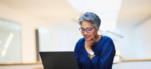Mulher idosa sorridente usando óculos e vestindo blusa azul escuro, olhando para laptop, em ambiente bem iluminado, expressando satisfação e felicidade.