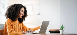 Mulher sorridente trabalhando no computador portátil em ambiente moderno com plantas e livros ao lado, representando produtividade e home office.