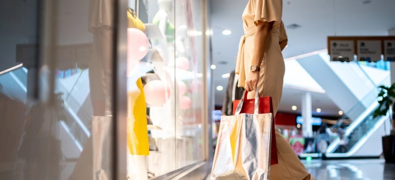 Mulher fazendo compras em shopping center, segurando sacolas de compras coloridas, com vitrines de lojas ao fundo. Cenário de comércio e moda.