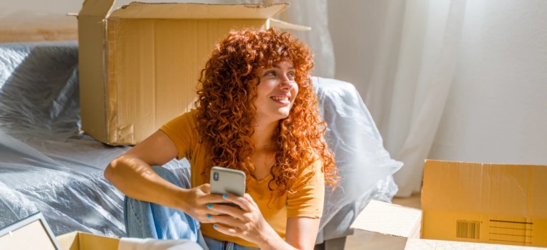 Mulher com cabelo cacheado vermelho sorrindo enquanto utiliza o celular em um ambiente de mudança, rodeada por caixas e móveis.
