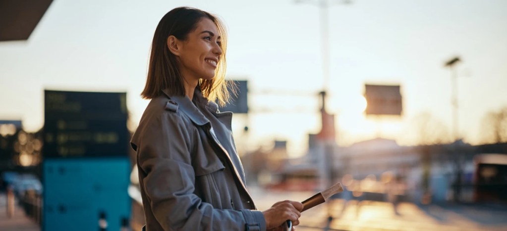 Mulher sorridente usando jaqueta em rua movimentada ao entardecer com luz natural.