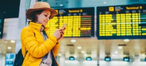 Mulher sorridente com chapéu, vestindo jaqueta amarela, usando o smartphone em balcão com informações de voos e terminal em aeroporto.