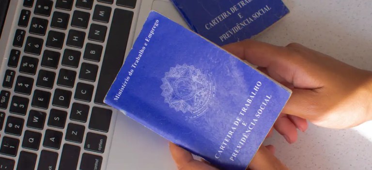 Pessoa segurando carteira de trabalho e previdência social ao lado de um notebook, destacando a importância desse documento para direitos trabalhistas no Brasil.