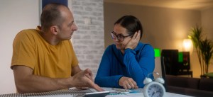 Homem e mulher conversando e discutindo questões financeiras ou pessoais enquanto sentados em uma mesa de jantar, com uma sala de estar ao fundo.