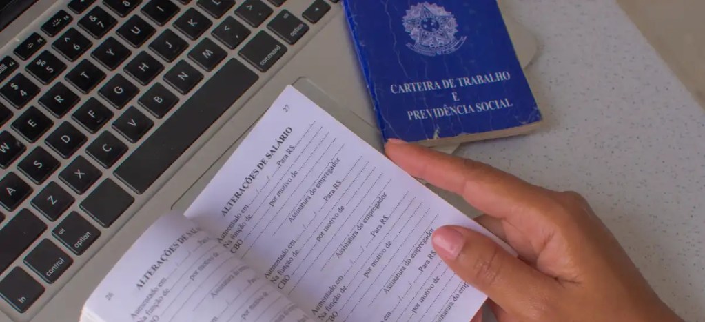Pessoa segurando um documento de inscrição de trabalho e previdência social ao lado de um laptop e carteira de trabalho azul, relacionados à busca de emprego.
