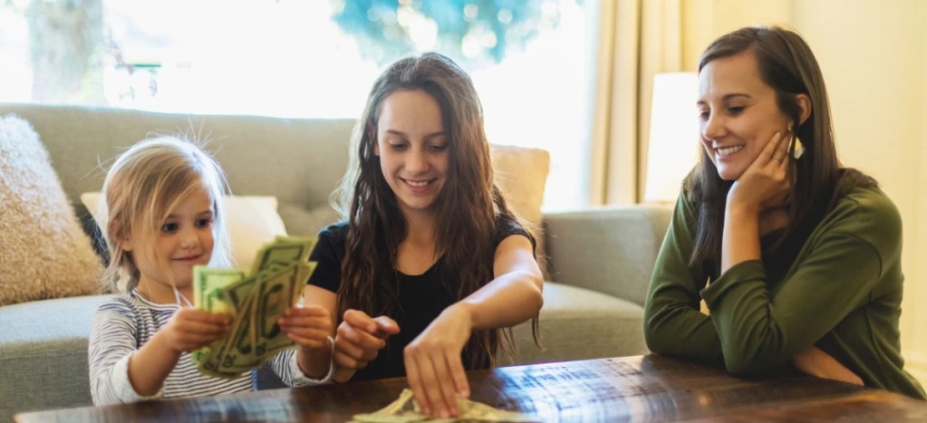 Família brincando e contando dinheiro em uma mesa na sala de estar, momento de diversão e economia com crianças e mãe
