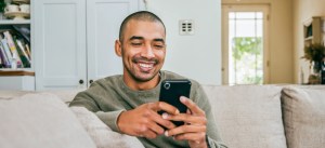 Homem sorridente usando celular enquanto está sentado no sofá em casa, ambiente aconchegante com livros e decoração moderna ao fundo.