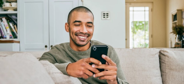 Homem sorridente usando celular enquanto está sentado no sofá em casa, ambiente aconchegante com livros e decoração moderna ao fundo.