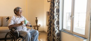 Idoso sentado em cadeira de rodas em casa, segurando uma xícara, ao lado de uma janela de vidro, ambiente acolhedor com cortina floral.
