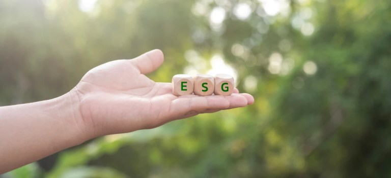 Mão segurando três dados com as letras E, S e G, simbolizando sustentabilidade, meio ambiente e responsabilidade social, com fundo natural e iluminação solar.