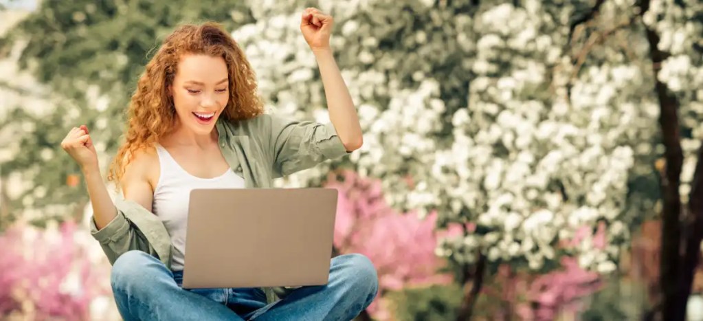 Jovem mulher feliz usando laptop ao ar livre na primavera, celebrando conquista no trabalho ou na vida pessoal em um parque com árvores floridas ao fundo.