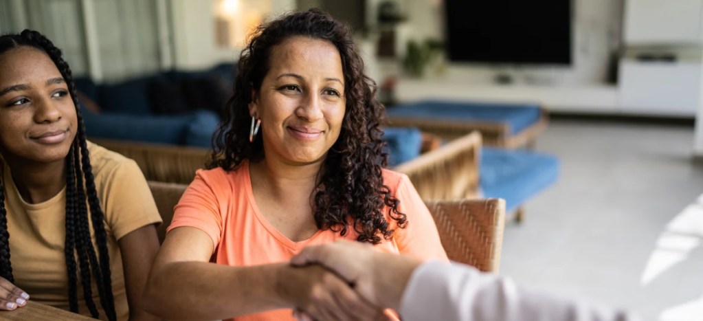 Mulher sorridente cumprimentando alguém em um ambiente acolhedor, com uma jovem ao lado, simbolizando confiança e conexão social.