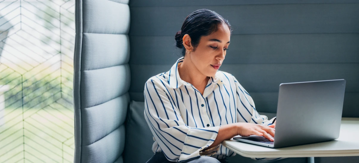 Mulher de cabelos pretos usando camisa listrada azul e branca, sentada em um ambiente moderno com sofá estofado, trabalhando em um laptop, com expressão concentrada.