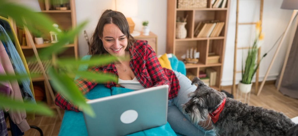 Mulher sorridente usando um laptop em casa com seu cachorro, ambiente acolhedor e bem decorado, ideal para quem busca conforto na casa.
