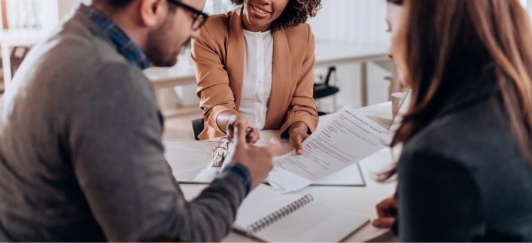Três pessoas em uma reunião de negócios, conversando e analisando documentos em um ambiente de escritório profissional, com sorriso e interação colaborativa.
