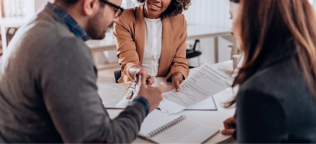Três pessoas em uma reunião de negócios, conversando e analisando documentos em um ambiente de escritório profissional, com sorriso e interação colaborativa.