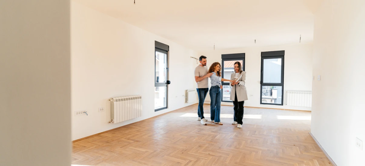 Três pessoas analisando um apartamento vazio com piso de madeira e janelas grandes, em uma visita imobiliária ou inspeção de residência.