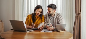 Casal trabalhando em home office: mulher escreve e faz anotações em um caderno com caneta, enquanto o homem usa o celular e analisa relatórios no laptop e tablet sobre a mesa, em sala com cortinas e luz natural.
