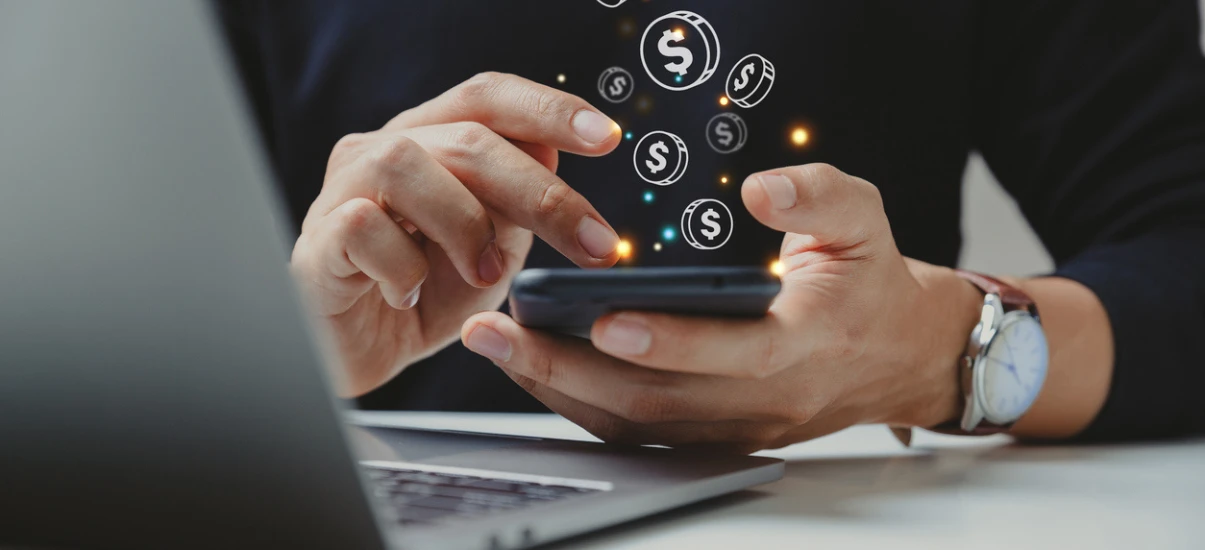 Pessoa manipulando smartphone com ícones de dólar e símbolos de dinheiro flutuando, representando finanças, investimentos ou dinheiro digital.