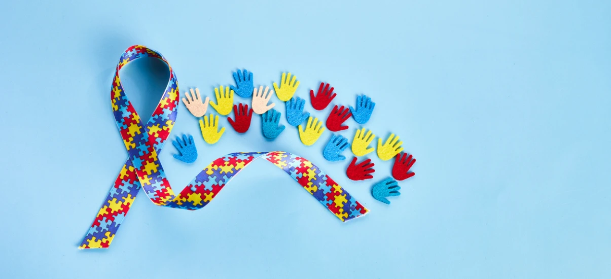 Laço de fita com padrão de quebra-cabeça, símbolo da conscientização sobre autismo, acompanhada por mãos de brinquedo coloridas azul, vermelho e amarelo, simbolizando apoio e inclusão em luta contra o autismo.