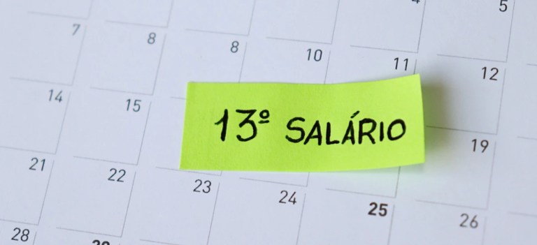 Anotação em papel amarelo neon com a frase “13º SALÁRIO” escrita à mão, destacada sobre um calendário com dias numerados ao fundo.