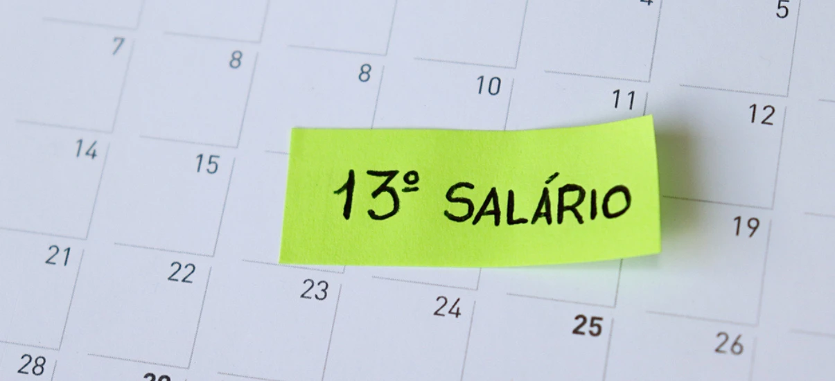 Anotação em papel amarelo neon com a frase “13º SALÁRIO” escrita à mão, destacada sobre um calendário com dias numerados ao fundo.