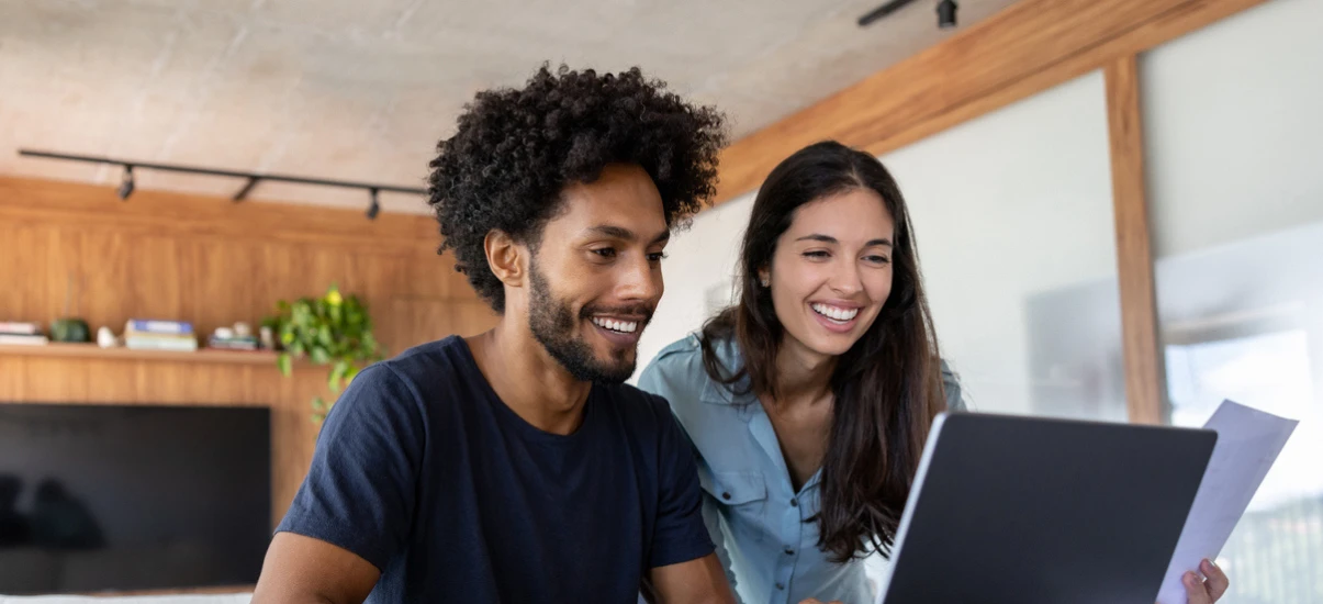 Casal sorridente em home office analisando informações em um laptop, com homem de cabelo crespo e mulher de camisa azul, ambiente moderno com madeira e plantas ao fundo.