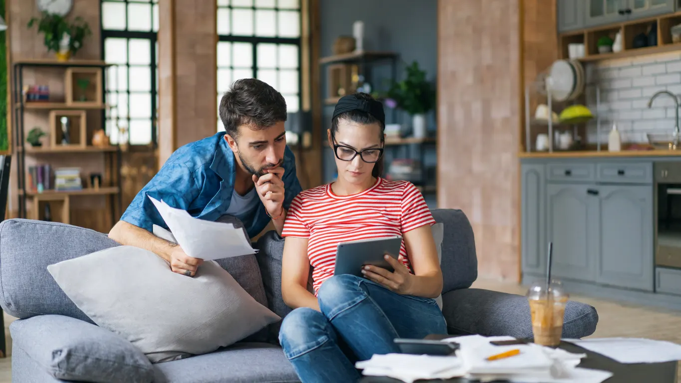 Jovens estudando e lendo documentos no sofá da sala, enquanto um deles usa um tablet; escritório com estantes e cozinha ao fundo, em ambiente moderno.