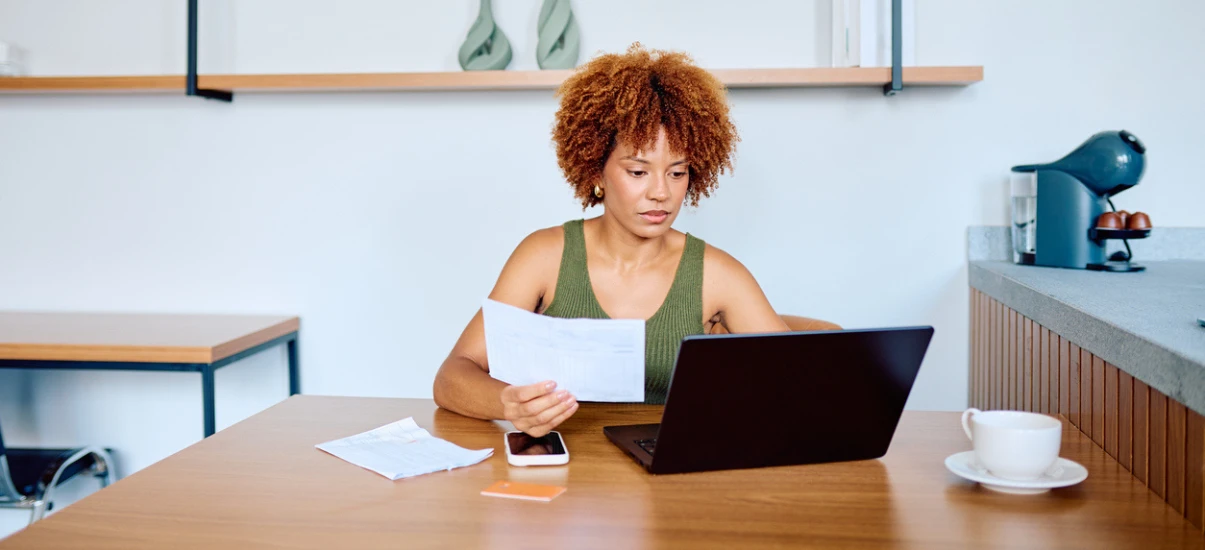 Mulher com cabelo cacheado em escritório contemporâneo, sentada à mesa com notebook aberto e lendo documentos em papel, com celular e xícara ao lado, ambiente de trabalho moderno e organizado.