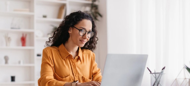 Mulher com cabelo cacheado e óculos, em um home office, trabalhando no notebook com postura tranquila e foco na tela, ambiente moderno e iluminado.