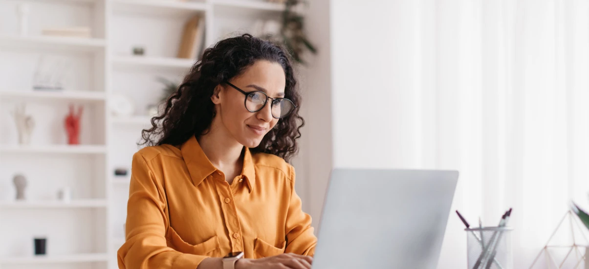 Mulher com cabelo cacheado e óculos, em um home office, trabalhando no notebook com postura tranquila e foco na tela, ambiente moderno e iluminado.