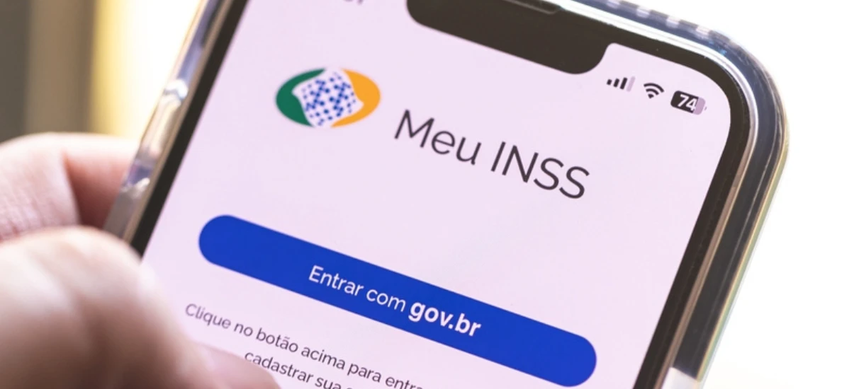 Celular na mão exibindo a tela de acesso ao Meu INSS com o botão “Entrar com gov.br”, ícone do serviço e indicador de sinal no canto superior direito.