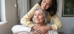 Mulher cuidando de idosa em casa, com carinho e apoio, segurando as mãos da senhora no sofá. Ambiente aconchegante e acolhedor.