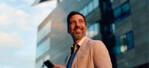 Homem sorridente com barba e cabelo escuro, usando terno claro e camisa social, segura um smartphone e caminha ao ar livre em frente a prédios modernos com janelas de vidro.