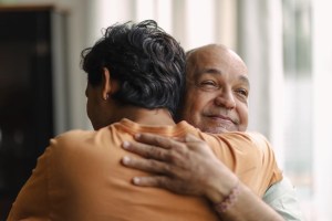 Homem abraçando um homem idoso carinhosamente em ambiente interno, transmitindo afeto e cuidado, com expressão de conforto e respeito.