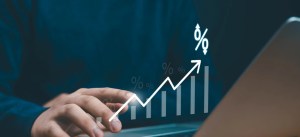 Mão apontando para gráfico de crescimento com setas e símbolos de porcentagem em fundo de escritório, sugerindo aumento de resultados e promoções