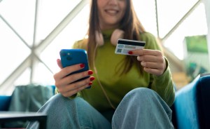 Mulher sentada segurando um cartão de crédito e um celular azul, em um ambiente iluminado, sugerindo pagamento por aplicativo e compras online.