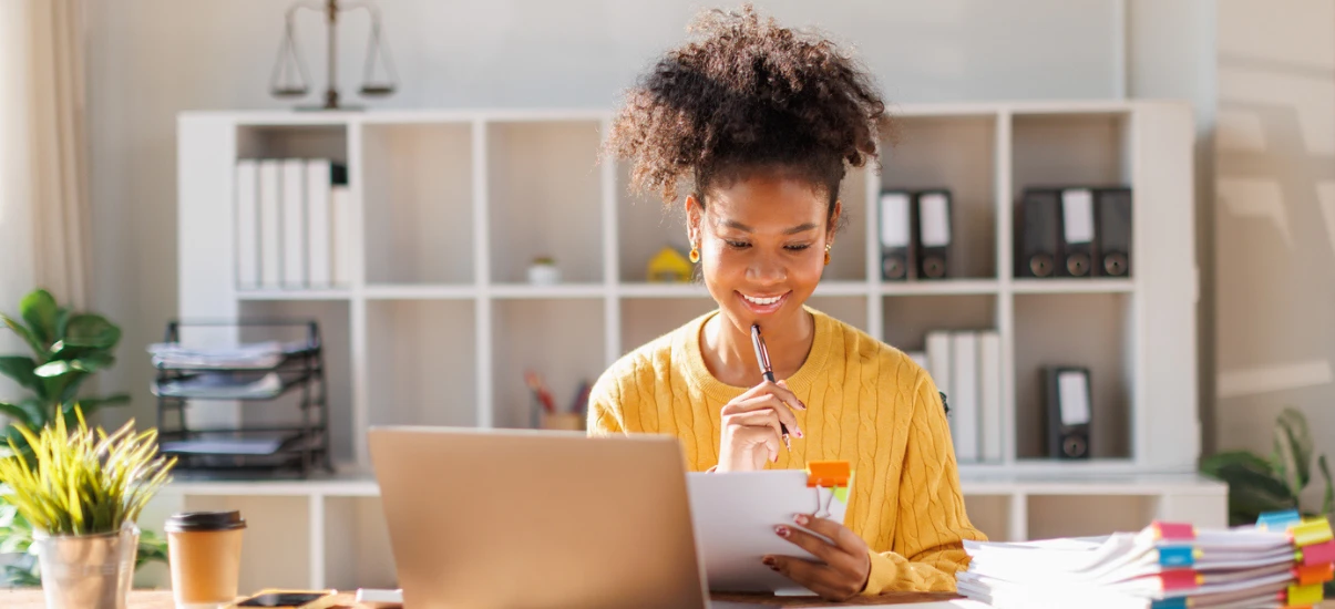 Mulher trabalhando em escritório moderno, sorridente, usando laptop e consultando documentos enquanto anota informações com caneta, ambiente organizado e iluminado.