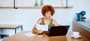Mulher com cabelo afro encaracolado trabalhando em uma mesa de escritório, analisando documentos e usando notebook, com xícara ao lado, em ambiente moderno.