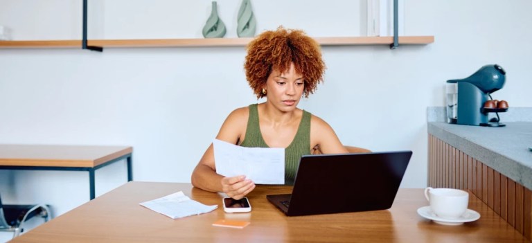 Mulher com cabelo afro encaracolado trabalhando em uma mesa de escritório, analisando documentos e usando notebook, com xícara ao lado, em ambiente moderno.