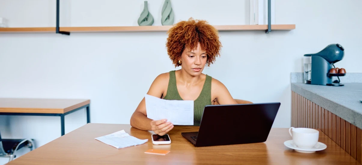Mulher com cabelo afro encaracolado trabalhando em uma mesa de escritório, analisando documentos e usando notebook, com xícara ao lado, em ambiente moderno.