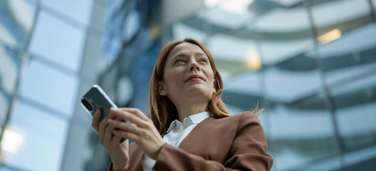 Mulher de negócios ao ar livre em ambiente corporativo, usando celular na mão enquanto olha para cima, com expressão confiante, fundo desfocado de prédios modernos